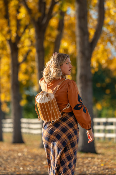 Woman in a plaid outfit with a brown leather jacket and backpack, standing in an autumn setting.